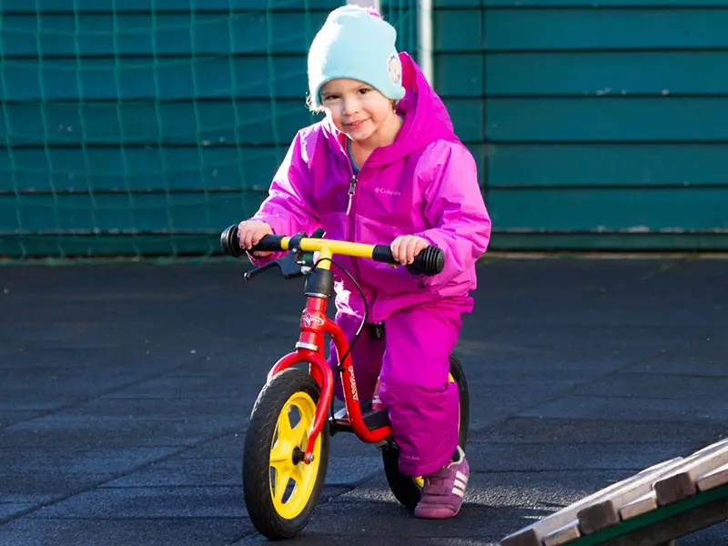 Girl in a pink snowsuit riding a balance bike on the outdoor court