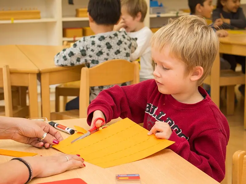 Boy practicing scissor cutting on paper with a teacher's guidance